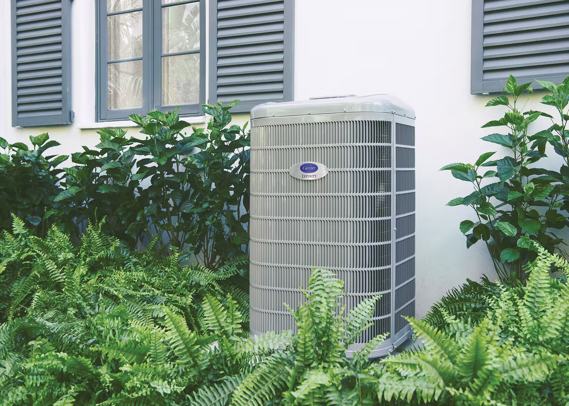 An outdoor air conditioning unit is installed next to a house, surrounded by green plants and located below a window with shutters.