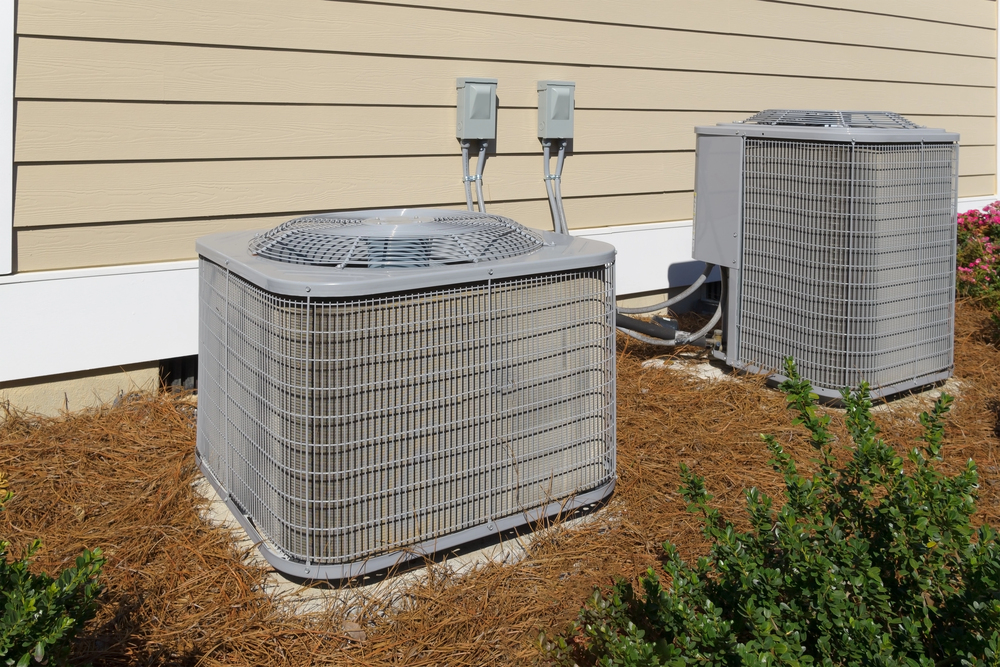 Two outdoor air conditioning units are installed on concrete pads next to a beige house with siding, surrounded by mulch and some green bushes.