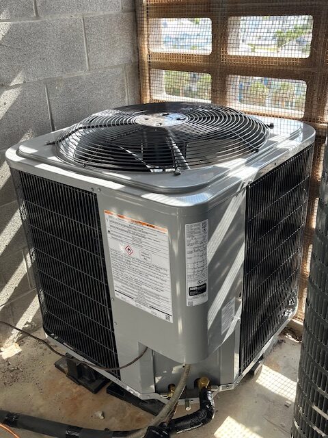 A central air conditioning unit is installed on a concrete floor near a cinder block wall and wire mesh window.