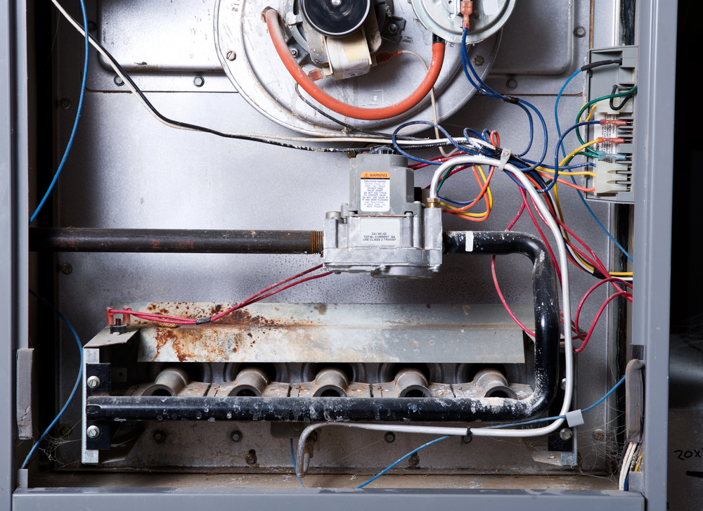 The interior of a gas furnace showing metal pipes, wires, a control valve, and burner tubes with some visible rust and dust buildup.
