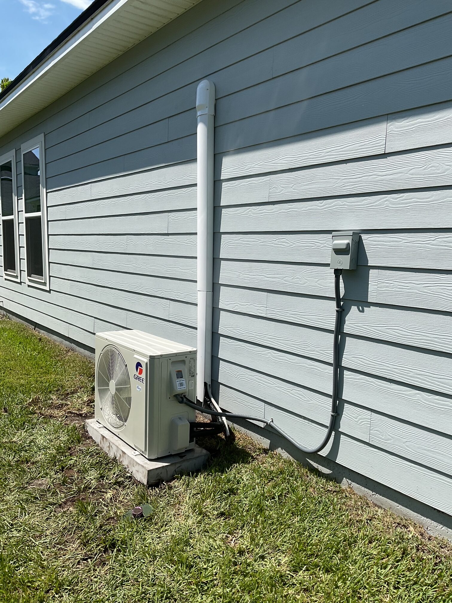 Outdoor air conditioning unit installed on a concrete pad next to the exterior wall of a light gray house, with pipes and electrical connections visible.