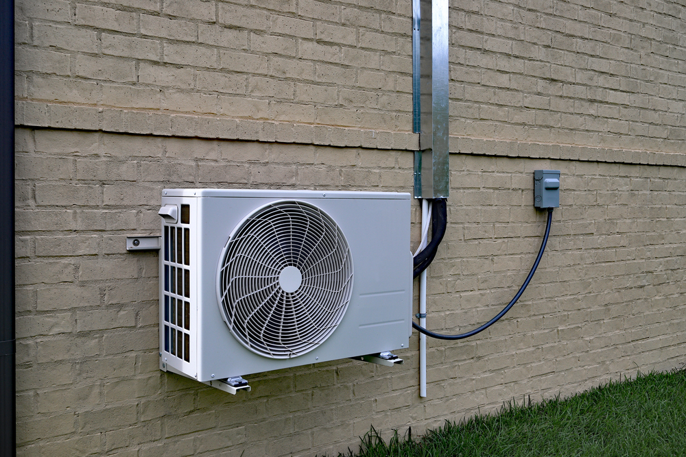 A white outdoor mini-split installation is mounted on a beige brick wall, with electrical wiring connected to a nearby utility box. Grass is visible at the bottom of the image.